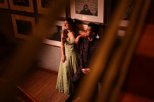 Load image into Gallery viewer, A couple in an intimate moment on a wooden staircase, viewed through a foreground blur. The woman wears a light sage green floral print maxi dress, and the man wears a dark patterned shirt.