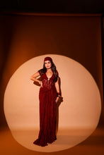 Load image into Gallery viewer, A woman modeling a floor-length maroon red gown with lace-up front detailing and a matching headpiece, standing inside a large circular spotlight on a warm brown studio backdrop.