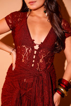 Load image into Gallery viewer, A close-up of a woman wearing a deep burgundy lace dress with a deep V-neckline, lace-up front, and a matching fabric tie belt at the waist. She is accessorized with red and gold bangles and dangling earrings against a warm beige background.