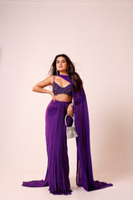 Load image into Gallery viewer, A woman models a purple two-piece traditional outfit featuring a heavily embellished lace crop top and a matching pleated skirt with a long draped dupatta, holding a small metallic handbag against a solid pale pink studio background.
