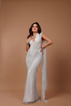 Load image into Gallery viewer, A woman modeling a white sleeveless gown with a deep V-neckline, front tie detail, and a long sheer draped overlay, posed against a solid warm tan studio backdrop.