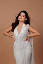 Load image into Gallery viewer, A woman posing in a white sleeveless halter-neck dress with a deep V-neckline, fringe details, and a draped sheer overlay, set against a solid warm brown studio background.