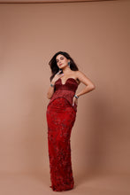 Load image into Gallery viewer, A woman modeling a strapless red evening gown with floral lace detailing, standing against a solid warm brown studio backdrop.