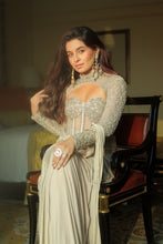 Load image into Gallery viewer, A woman seated on a dark wooden chair with red upholstery, wearing an embellished cream and silver long-sleeve gown with a sheer bodice and draped skirt. She is wearing large dangling earrings and rings in an indoor setting.