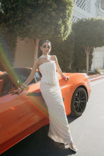 Load image into Gallery viewer, A woman wearing a strapless white corset-style maxi dress and sunglasses poses next to an orange sports car on a sunlit street with trees and a building in the background.

