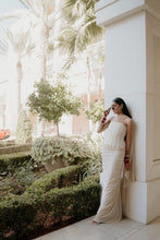 Load image into Gallery viewer, A woman wearing a strapless cream-colored gown leans against a white architectural column in a sunlit outdoor setting with palm trees and manicured hedges.
