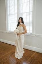Load image into Gallery viewer, A woman stands indoors in a bright room with white walls, window blinds, and hardwood floors, wearing a strapless ivory satin gown and holding a bouquet of white flowers.
