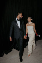 Load image into Gallery viewer, A couple walking hand-in-hand backstage against a black curtain. The man is wearing a classic black tuxedo with a white shirt and black bow tie, and the woman is wearing a strapless cream-colored gown with red gloves.
