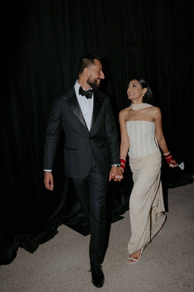 A couple walking hand-in-hand backstage against a black curtain. The man is wearing a classic black tuxedo with a white shirt and black bow tie, and the woman is wearing a strapless cream-colored gown with red gloves.