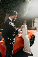 Load image into Gallery viewer, A bride in a strapless white wedding gown and a groom in a black tuxedo stand beside an orange convertible car outdoors, with sunlight and trees in the background.
