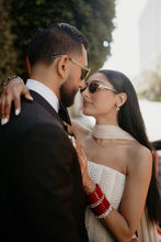 Load image into Gallery viewer, A couple in an intimate moment outdoors. The woman wears a strapless cream beaded dress, red stacked bangles, and gold-framed sunglasses, while the man is in a black suit jacket and white shirt.
