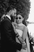 Load image into Gallery viewer, A black and white lifestyle photograph of a couple embracing outdoors. The woman wears a strapless textured white dress and dark sunglasses, while the man is in a dark suit and sunglasses.
