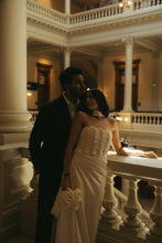 Load image into Gallery viewer, A couple posing on an ornate indoor staircase. The woman wears a strapless cream wedding gown with a textured bodice and holds a matching cream floral bouquet.
