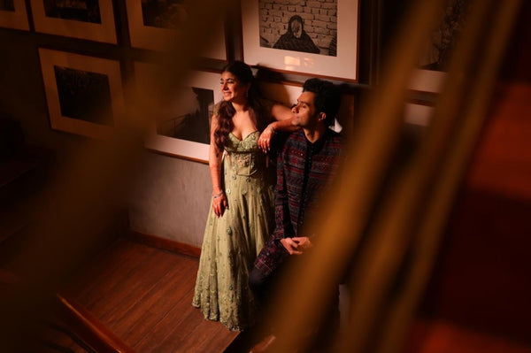 A couple in an intimate moment on a wooden staircase, viewed through a foreground blur. The woman wears a light sage green floral print maxi dress, and the man wears a dark patterned shirt.