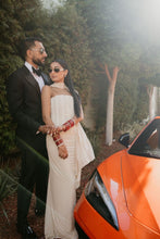 Load image into Gallery viewer, A couple standing outdoors next to an orange sports car, with the woman wearing a strapless cream-colored dress and red bangles, and the man wearing a black suit.
