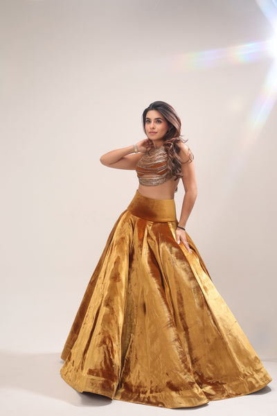A woman modeling a traditional Indian outfit consisting of a metallic gold velvet skirt and a matching embellished crop top, set against a light gray studio background with a lens flare effect.