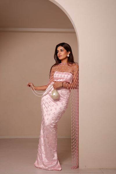 A woman models a light pink evening gown with a sheer mesh overlay draped over her shoulders, standing in a minimalist beige room with an arched doorway.
