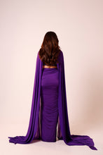 Load image into Gallery viewer, A woman viewed from behind wearing a floor-length purple gown with a long draped cape-like overlay, posed against a plain light beige studio background.
