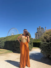 Load image into Gallery viewer, A woman wearing a strapless orange and gold evening gown stands outdoors on a garden path with hedges, a large Ferris wheel, and a historic building in the background under a clear blue sky.
