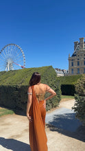 Load image into Gallery viewer, A woman stands outdoors in a garden setting wearing a sleeveless rust-orange draped evening gown with a gold embellished back panel. The background features a large green hedge, a blue sky, a ferris wheel, and a historic building.
