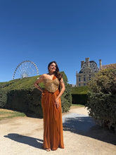 Load image into Gallery viewer, A woman posing outdoors in a long, floor-length rust-orange gown with a gold beaded bodice, set against a backdrop of manicured hedges, a castle, and a ferris wheel under a clear blue sky.
