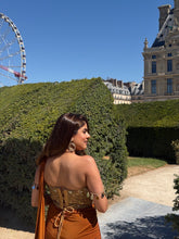Load image into Gallery viewer, A woman wearing a brown strapless corset-style dress with gold sequin detailing and a matching brown bag, standing outdoors in front of a large green hedge and a building under a clear blue sky.
