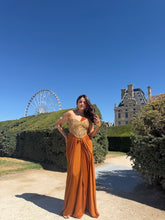 Load image into Gallery viewer, A woman wearing a sleeveless rust-orange maxi dress with a gold sequined bodice stands outdoors on a gravel path, with manicured hedges, a large building, and a Ferris wheel in the background under a clear blue sky.
