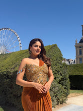 Load image into Gallery viewer, A woman wearing a sleeveless, off-the-shoulder rust-orange and gold embellished gown stands outdoors in front of green hedges and a blue sky, with a Ferris wheel and building visible in the background.
