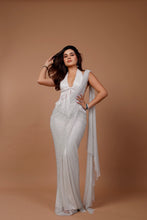 Load image into Gallery viewer, A woman modeling a white sleeveless maxi dress with a deep V-neckline, front fringe detailing, and a draped sheer overlay, set against a solid warm brown studio backdrop.
