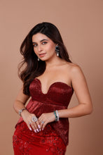 Load image into Gallery viewer, A woman posing in a strapless, deep red sequined gown with a sweetheart neckline and a red embroidered skirt, accessorized with silver and green statement jewelry against a solid tan background.
