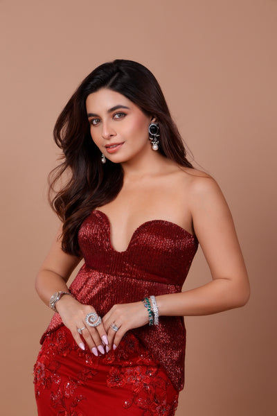 A woman posing in a strapless, deep red sequined gown with a sweetheart neckline and a red embroidered skirt, accessorized with silver and green statement jewelry against a solid tan background.
