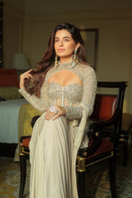 Load image into Gallery viewer, A woman posing in an elegant, heavily embellished cream-colored traditional gown with a sweetheart neckline and long sleeves, seated on a dark wooden chair in a warmly lit room.
