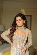 Load image into Gallery viewer, A woman wearing a beige and silver embellished long-sleeve top with a sheer corset-style bodice, paired with a draped cream saree, standing in a softly lit indoor setting.
