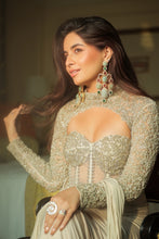 Load image into Gallery viewer, A woman wearing an ornate beige embellished long-sleeve dress with a sweetheart neckline and matching draped fabric, styled with large statement earrings and rings in an indoor setting.
