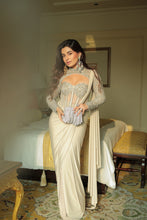 Load image into Gallery viewer, A woman stands in a bedroom wearing a cream-colored saree with heavy silver embroidery on the bodice and sleeves, holding a small silver clutch.
