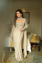 Load image into Gallery viewer, A woman models a cream-colored embellished saree with a matching sheer embroidered blouse in a decorated bedroom setting.
