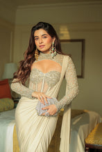 Load image into Gallery viewer, A woman wearing a heavily embellished cream and gold saree with long sheer embroidered sleeves, standing in a warmly lit bedroom setting.
