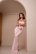 Load image into Gallery viewer, A woman models a light pink embellished saree with a sheer mesh top and a long matching draped scarf, standing in a minimalist beige room with an arched doorway.
