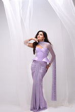 Load image into Gallery viewer, A woman models a lavender purple traditional Indian outfit consisting of a fitted corset-style top, matching draped skirt, and sheer long sleeves, posed against a white studio backdrop with hanging sheer fabric.
