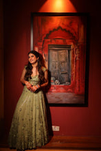 Load image into Gallery viewer, A woman in a floor-length olive green embellished gown stands in a dimly lit room beside a large framed painting of a rustic wooden door set in a red archway.
