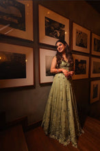Load image into Gallery viewer, A woman wearing a sleeveless olive green evening gown with intricate floral embroidery and beadwork, standing in a dimly lit room with framed black-and-white photographs on a dark wall.
