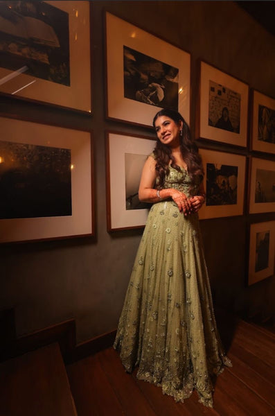 A woman wearing a sleeveless olive green evening gown with intricate floral embroidery and beadwork, standing in a dimly lit room with framed black-and-white photographs on a dark wall.
