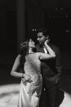 Load image into Gallery viewer, A black and white photograph of a couple embracing in a dimly lit indoor setting. The woman wears a strapless embellished gown and holds a matching handbag, while the man wears a dark suit.

