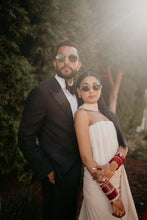 Load image into Gallery viewer, A couple posing outdoors in formal wear. The man wears a black tuxedo with a white shirt and black bow tie, and the woman wears a strapless cream-colored dress with red bangles.
